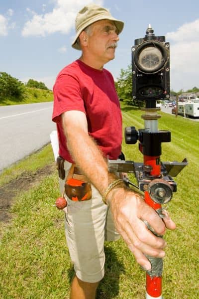 Professional land surveyor using equipment in the field beside a road and grassy area to confirm property boundaries